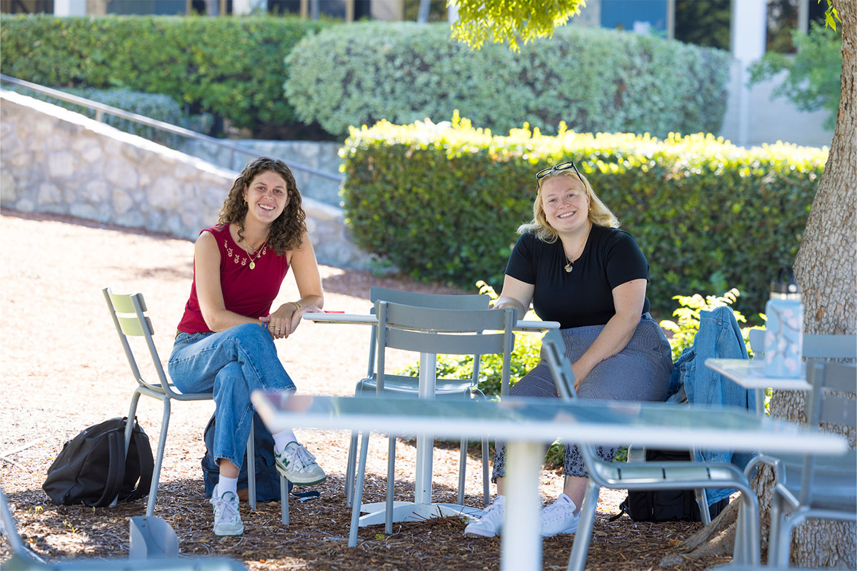 Two female students seated at a table under a tree.