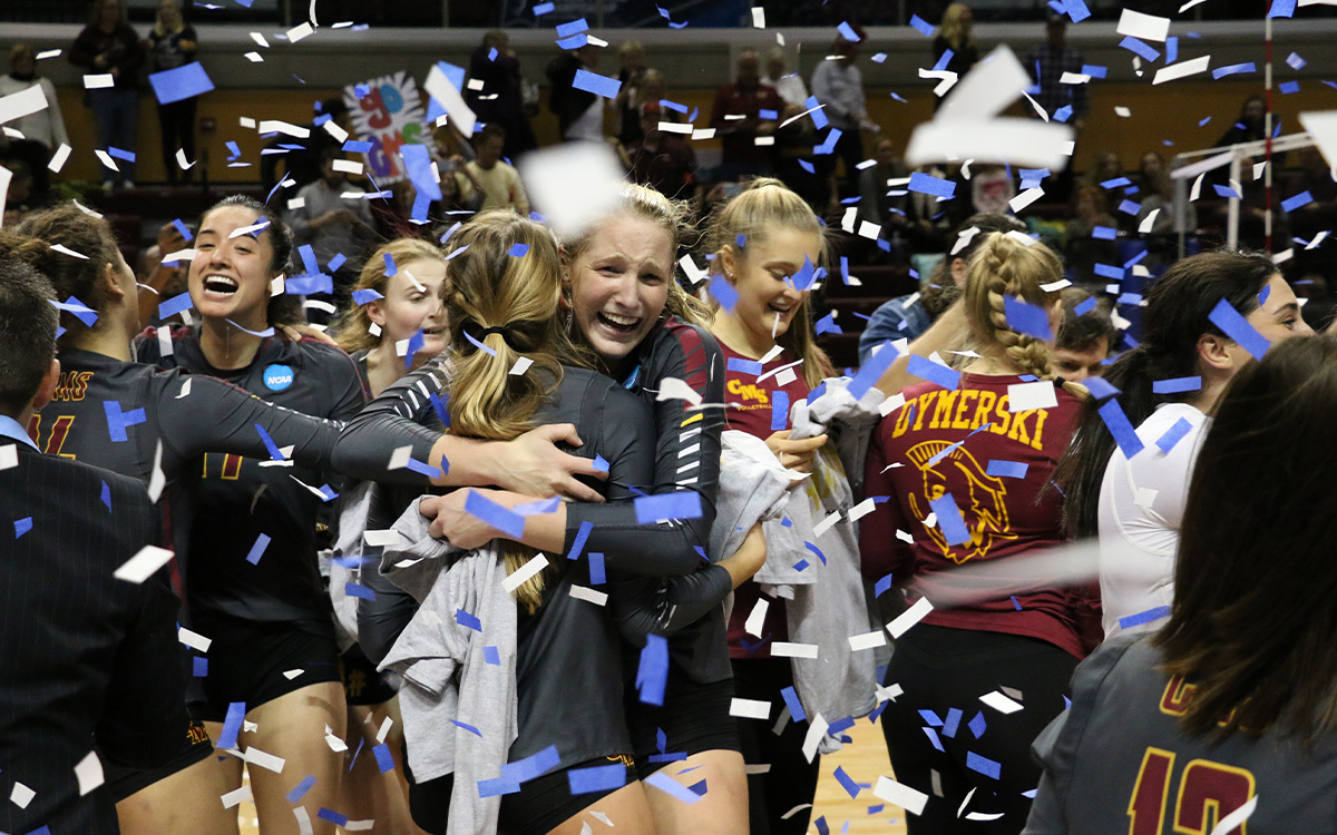 Two volleyball players hug in the foreground while the rest of the team celebrates their championship win.