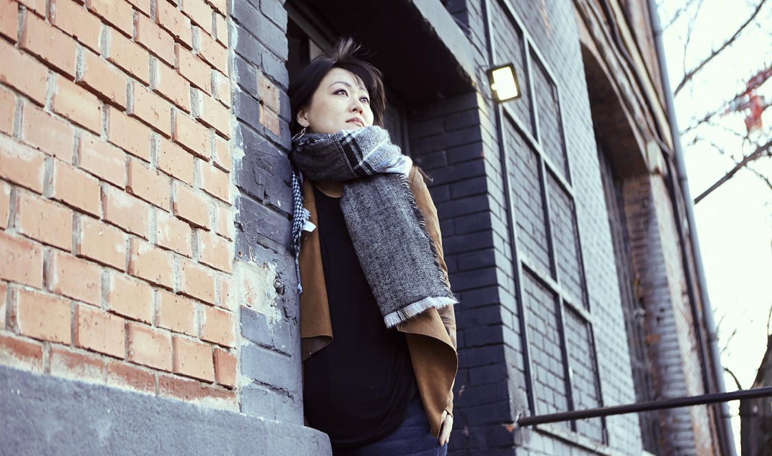 A photo of alumna Tracy Wang '04 leans against a building that's color coordinated with her cold weather outfit. A bare tree in the background indicates fall season.