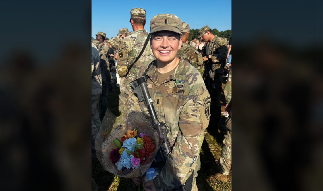 Lauren Jeans '24 in military fatigues holds a rifle and bouquet of flowers.