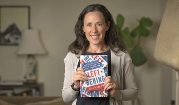 Professor Lily Geismer photographed at home, smiling and holding up her latest book, &quot;Left Behind.&quot;