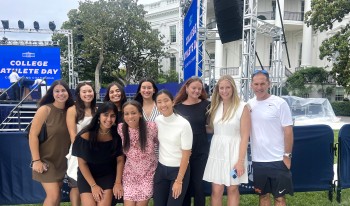 Group of female CMC tennis players and their coach stand and squat in front of a blue stage set up next to the White House for the event.