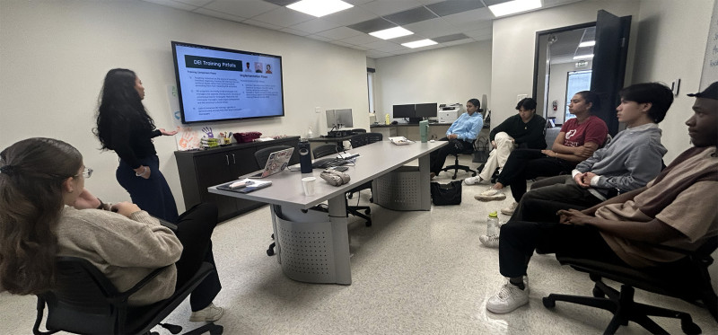 Students seated in a semi-circle facing a projector while a professor leads discussion.
