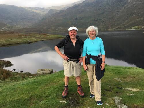 Man wearing black top and brown bottoms next to woman in teal and brown bottoms standing in front of mountains with lake.