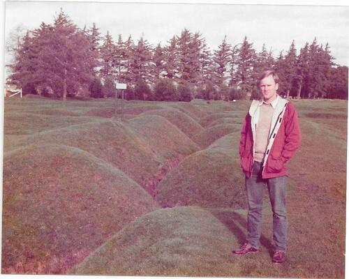 Man standing on grassy hills wearing red sweater and jeans. 